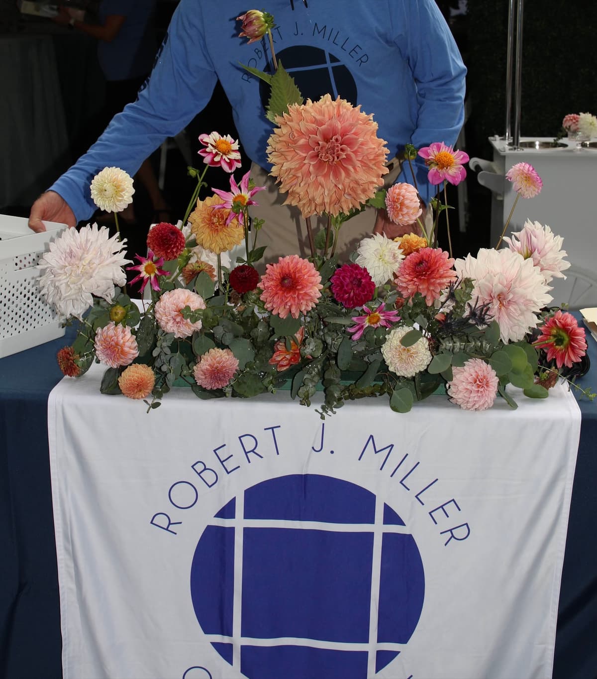 A lush floral centerpiece featuring a massive peach dinnerplate dahlia surrounded by smaller dahlias in shades of white, burgundy, and coral, arranged with eucalyptus greenery on a table with a blue and white logo cloth.