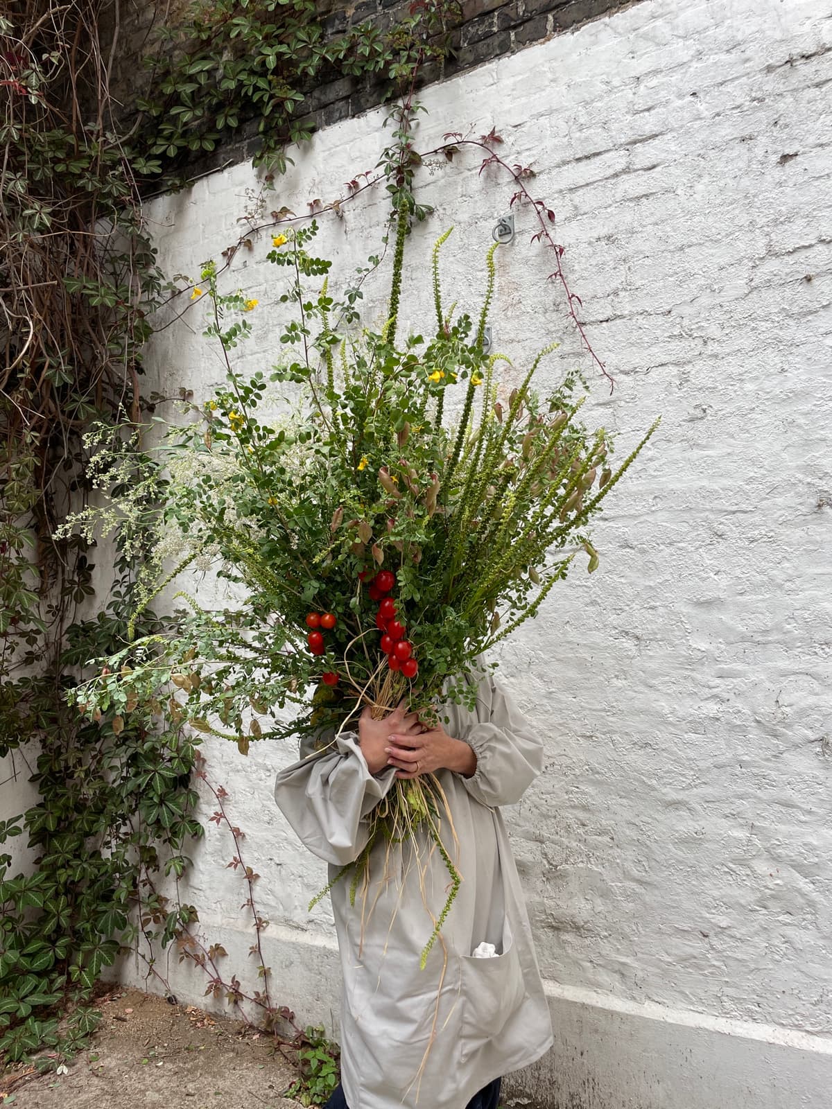 A florist wearing a beige linen dress holds a massive, overflowing armful of wild greenery, tall grasses, and clusters of red cherry tomatoes on the vine against a textured white brick wall.