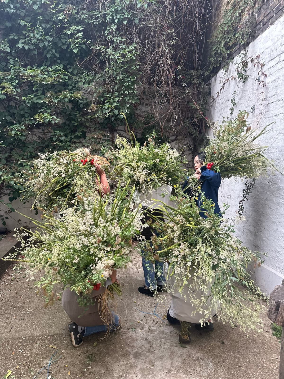 Three florists standing outdoors against a weathered white brick wall, holding massive, overflowing bundles of white and green wildflowers, grasses, and amaranth with small red berry accents.