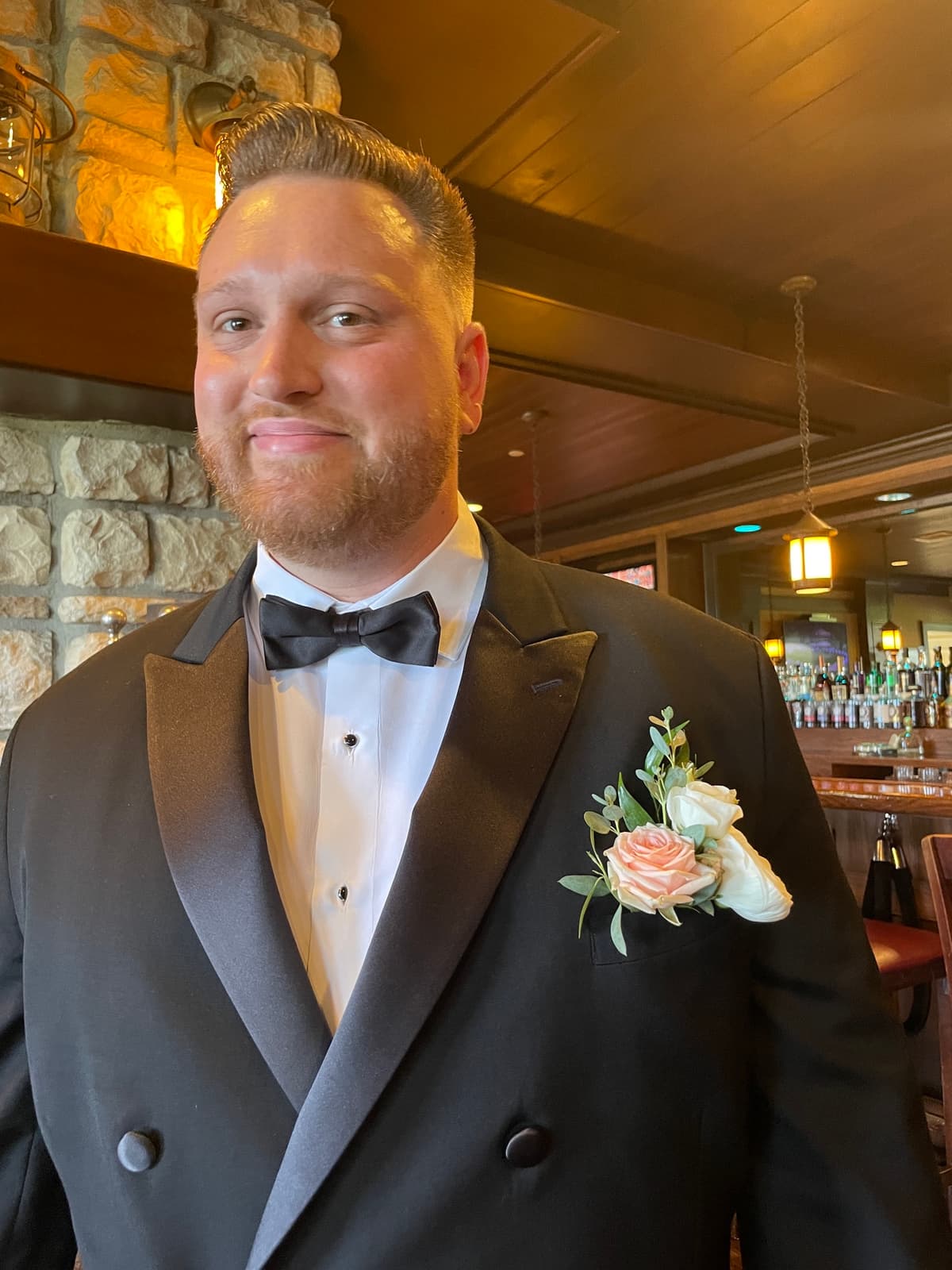 A smiling groom wearing a black tuxedo with a boutonniere featuring white and blush spray roses accented with eucalyptus greenery.