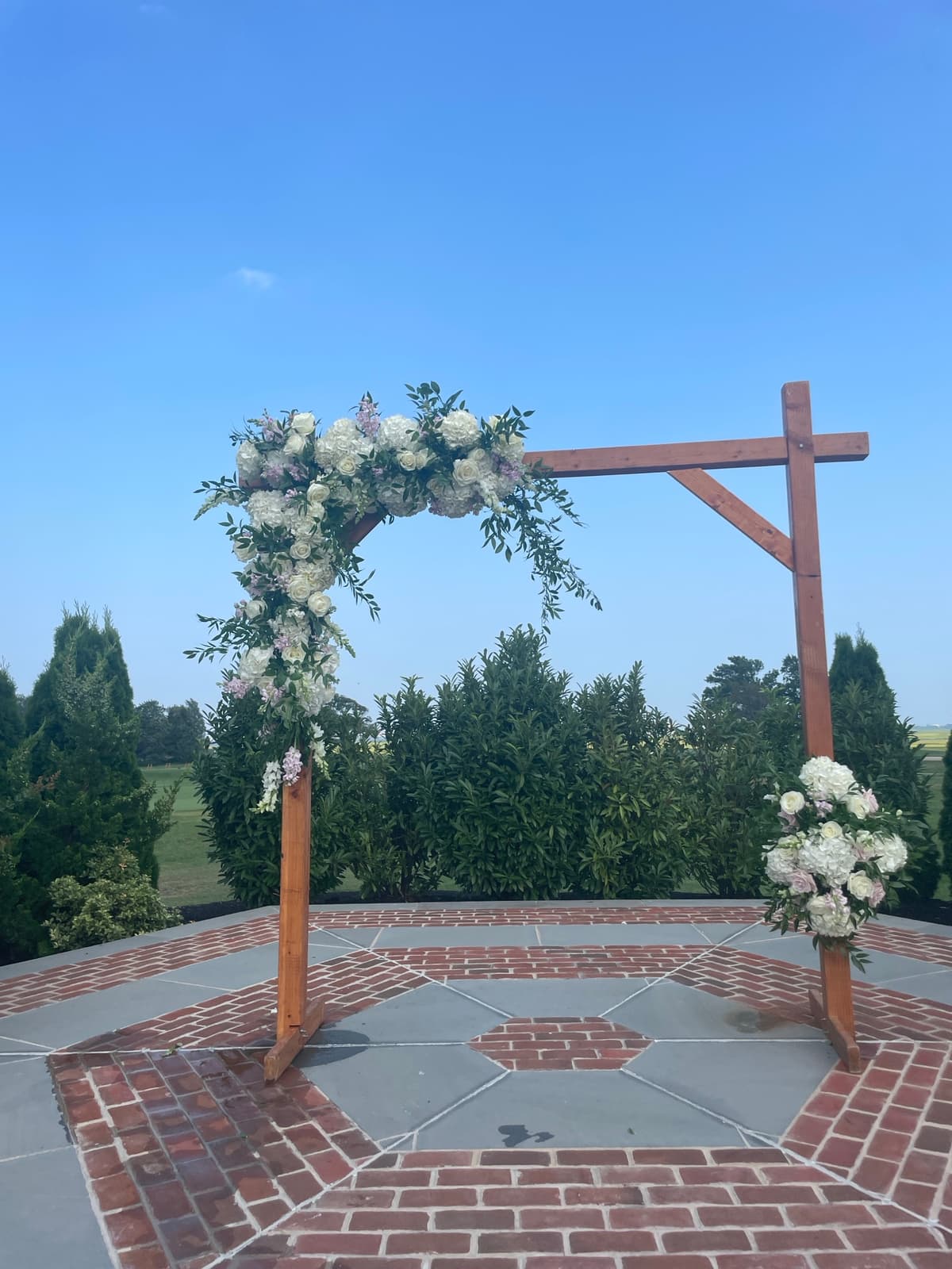 A wooden wedding arch standing on a brick and stone patio against a clear blue sky, decorated with an asymmetrical arrangement of white hydrangeas, cream and blush roses, and trailing greenery.
