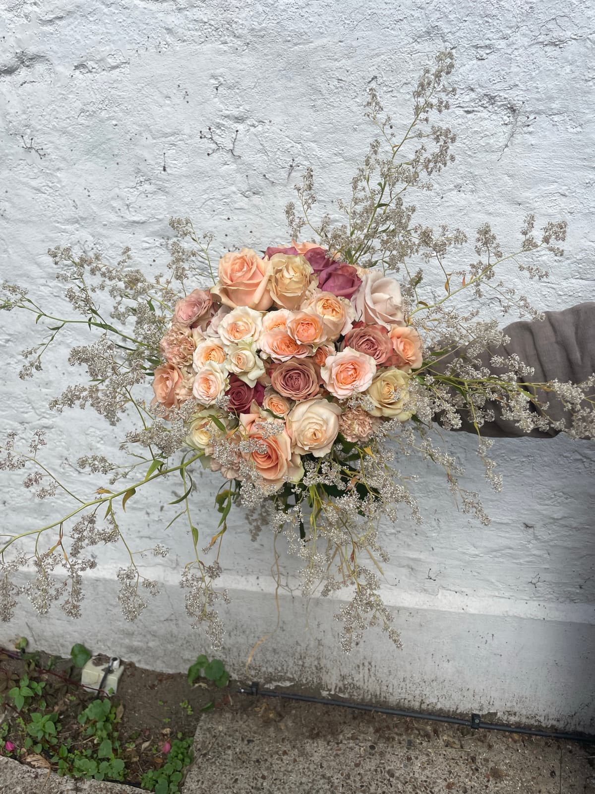 A top-down view of a lush bouquet held against a white textured wall, featuring roses in shades of peach, apricot, blush, and dusty mauve surrounded by a halo of airy white dried filler flowers.