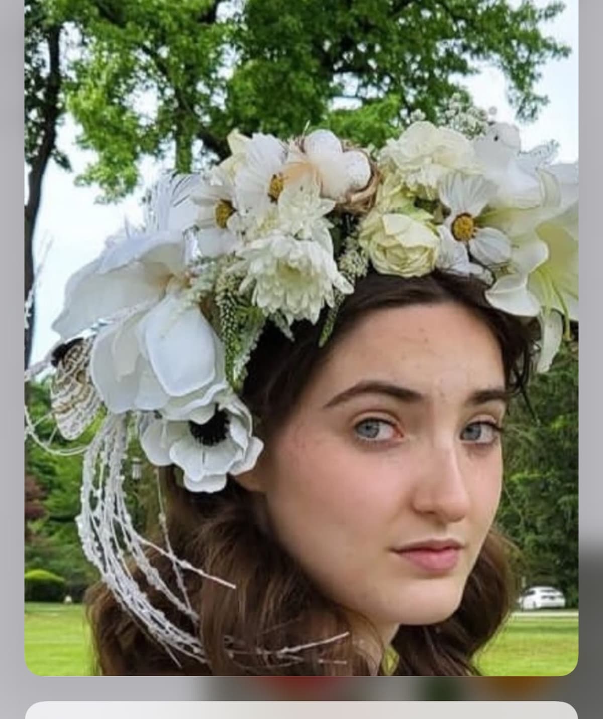 A young woman wearing an elaborate white and cream floral crown featuring anemones, roses, and trailing lace against a lush green outdoor backdrop.