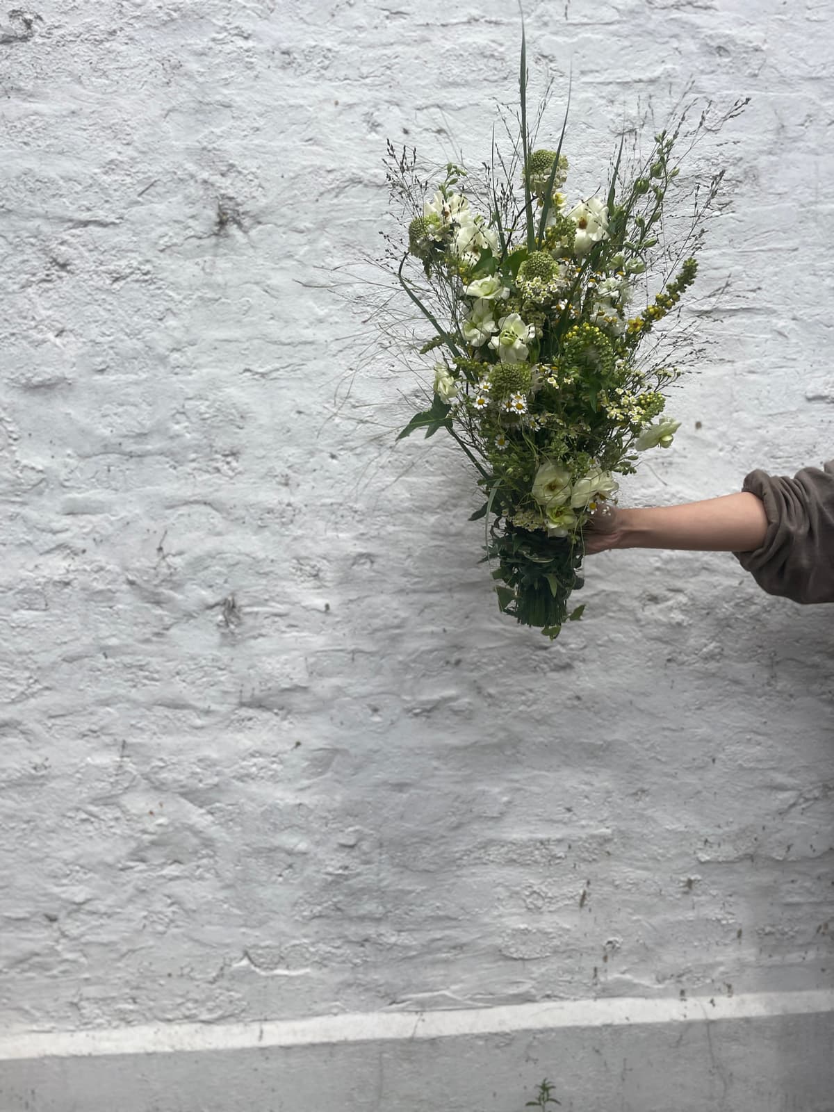 A hand holding a lush, meadow-inspired bouquet featuring white sweet peas, feverfew, and tall wispy grasses against a textured white brick wall.
