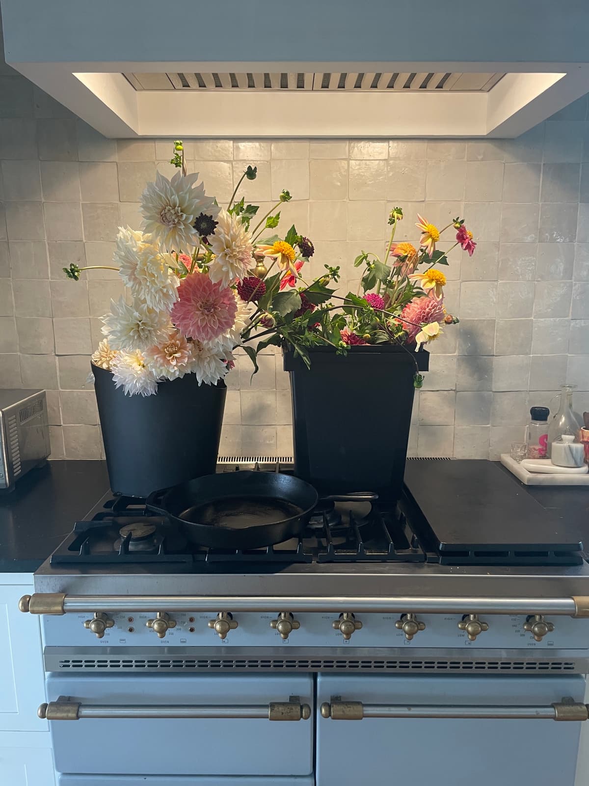 A collection of fresh dahlia flowers in black harvest buckets sitting on a blue kitchen range with brass knobs, set against a white tiled backsplash.