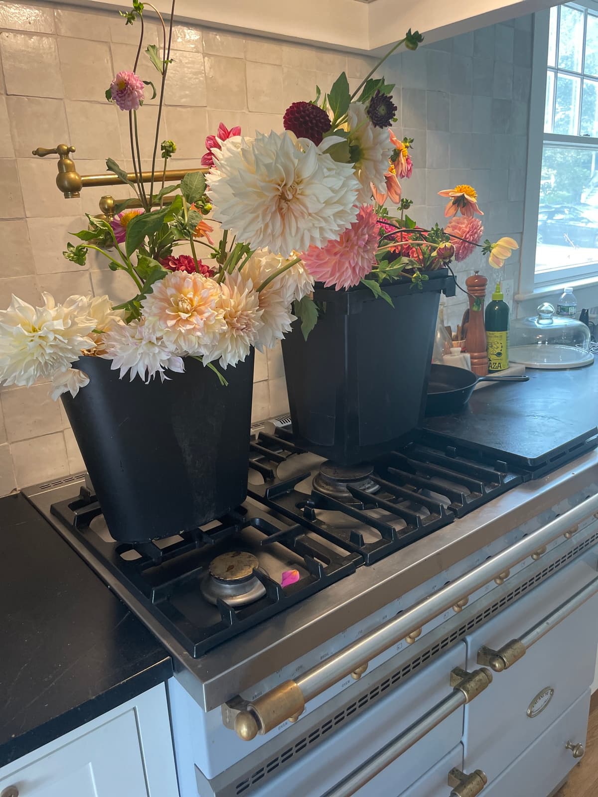 Black buckets filled with fresh dahlia flowers resting on a high-end kitchen range with brass knobs, illuminated by natural window light.