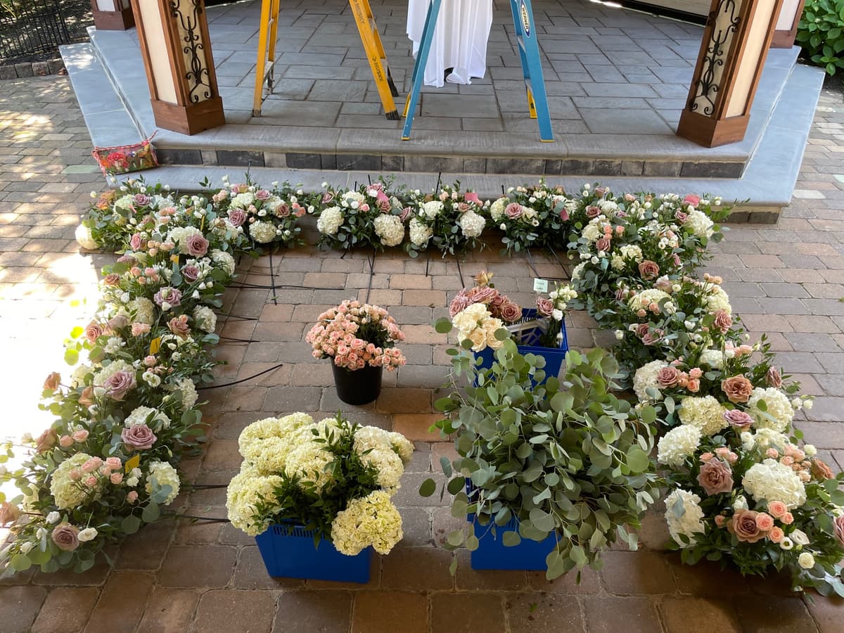 High-angle shot of a floral wedding setup in progress on a paved patio, featuring a lush border of white hydrangeas and mauve roses, with buckets of eucalyptus and spray roses in the center.