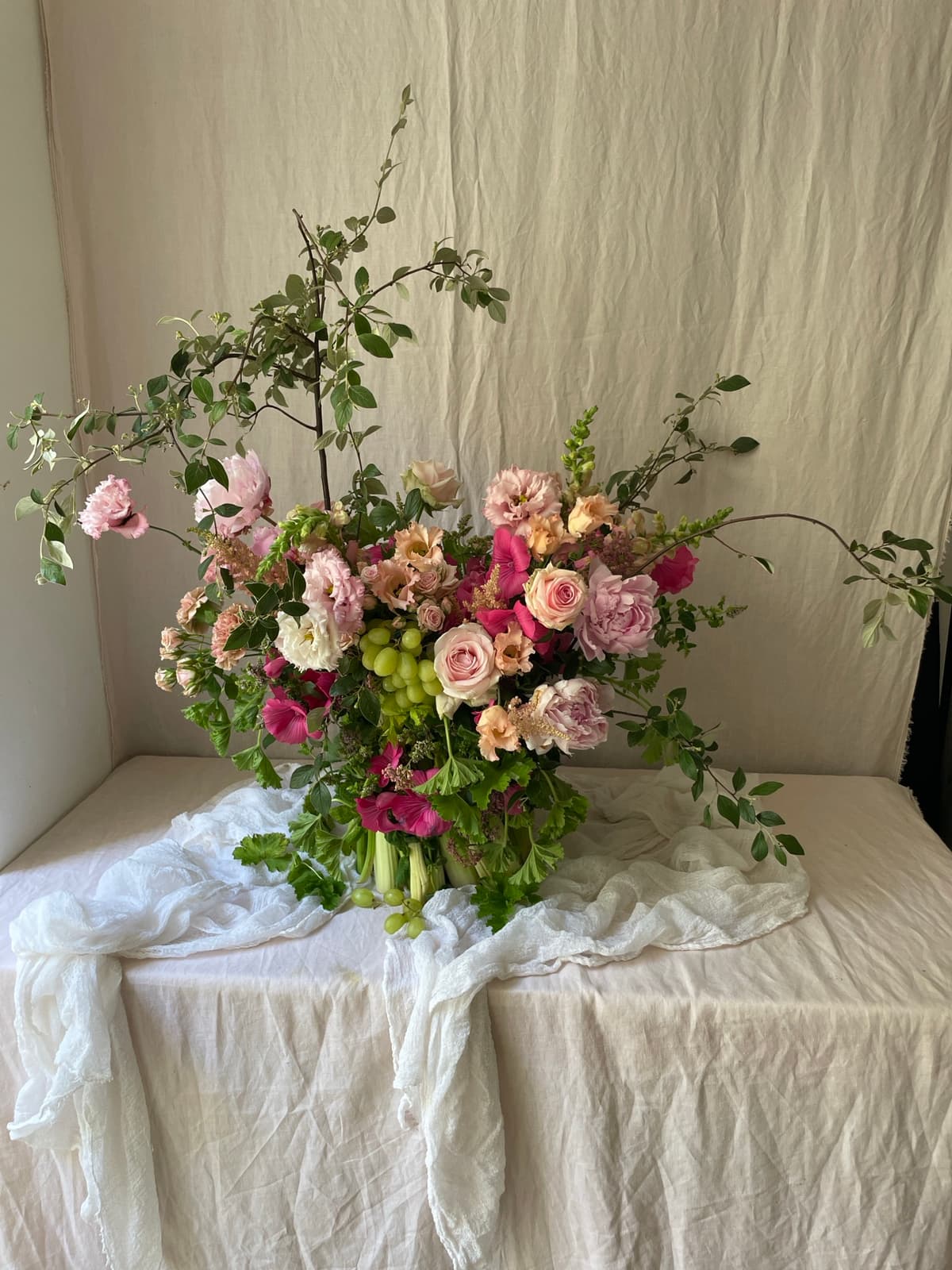A lush, asymmetrical floral arrangement featuring blush roses, pink lisianthus, and bright magenta blooms with clusters of green grapes, set against a draped beige linen background.