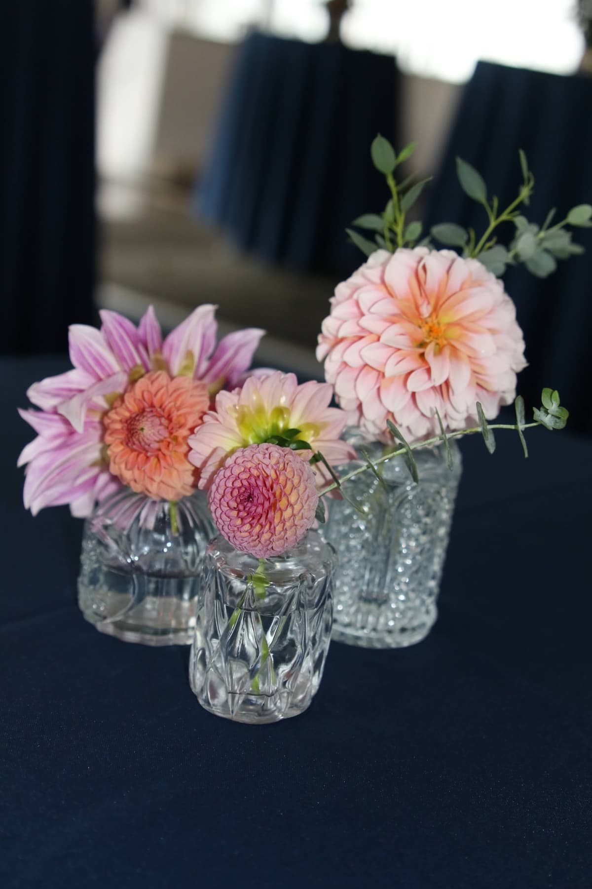 Three vintage cut-glass bud vases arranged on a dark blue tablecloth, filled with pink, peach, and lavender dahlias and sprigs of eucalyptus.