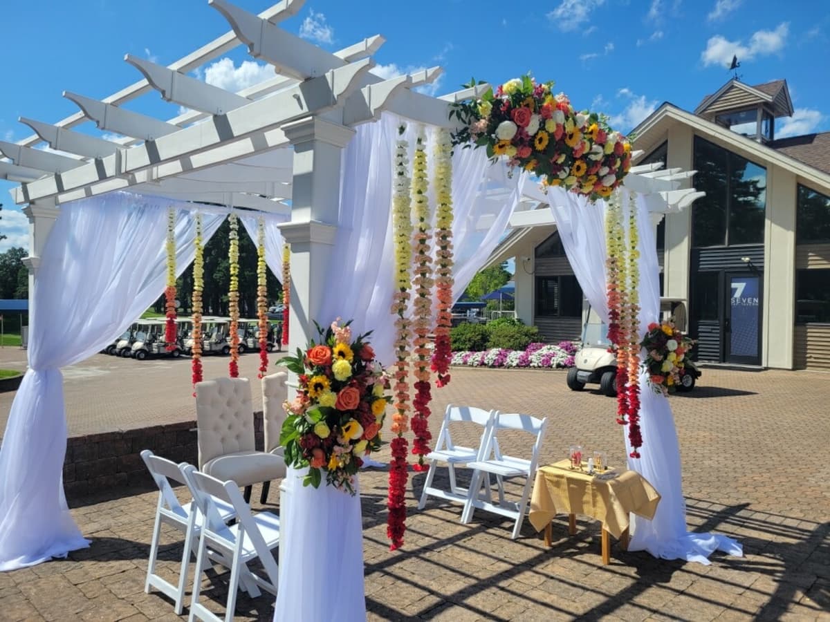 A sunny outdoor wedding ceremony setup featuring a white pergola draped in sheer white fabric. The focal point is a backdrop of hanging floral garlands in an ombre gradient, transitioning from yellow to orange to deep red. Matching floral arrangements with sunflowers and roses adorn the pergola posts.