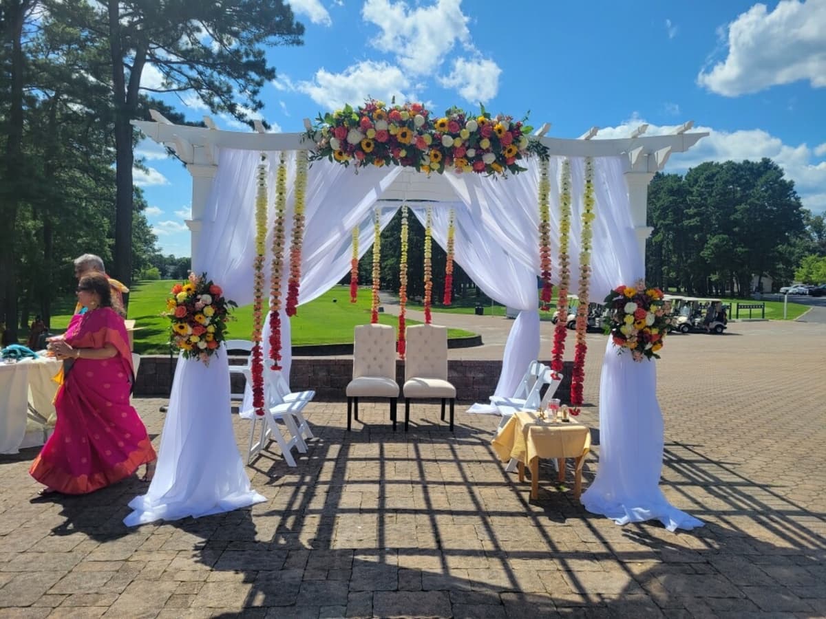 Outdoor wedding mandap with a white pergola draped in sheer white fabric, featuring a top floral arrangement of sunflowers and roses, matching side bouquets, and hanging ombre marigold garlands in yellow and red.