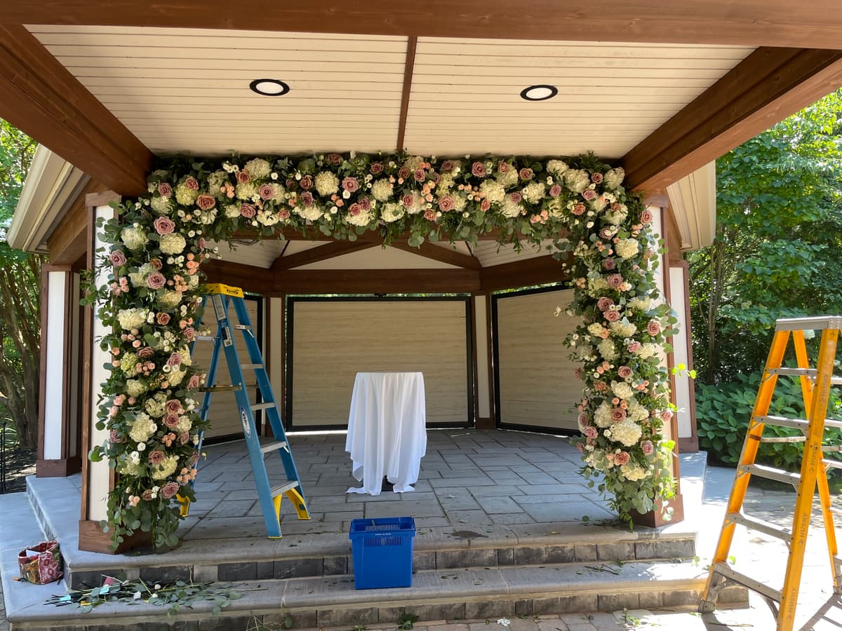 A lush floral archway installation on a wooden gazebo featuring white hydrangeas, dusty pink roses, and eucalyptus greenery, with ladders visible during setup.