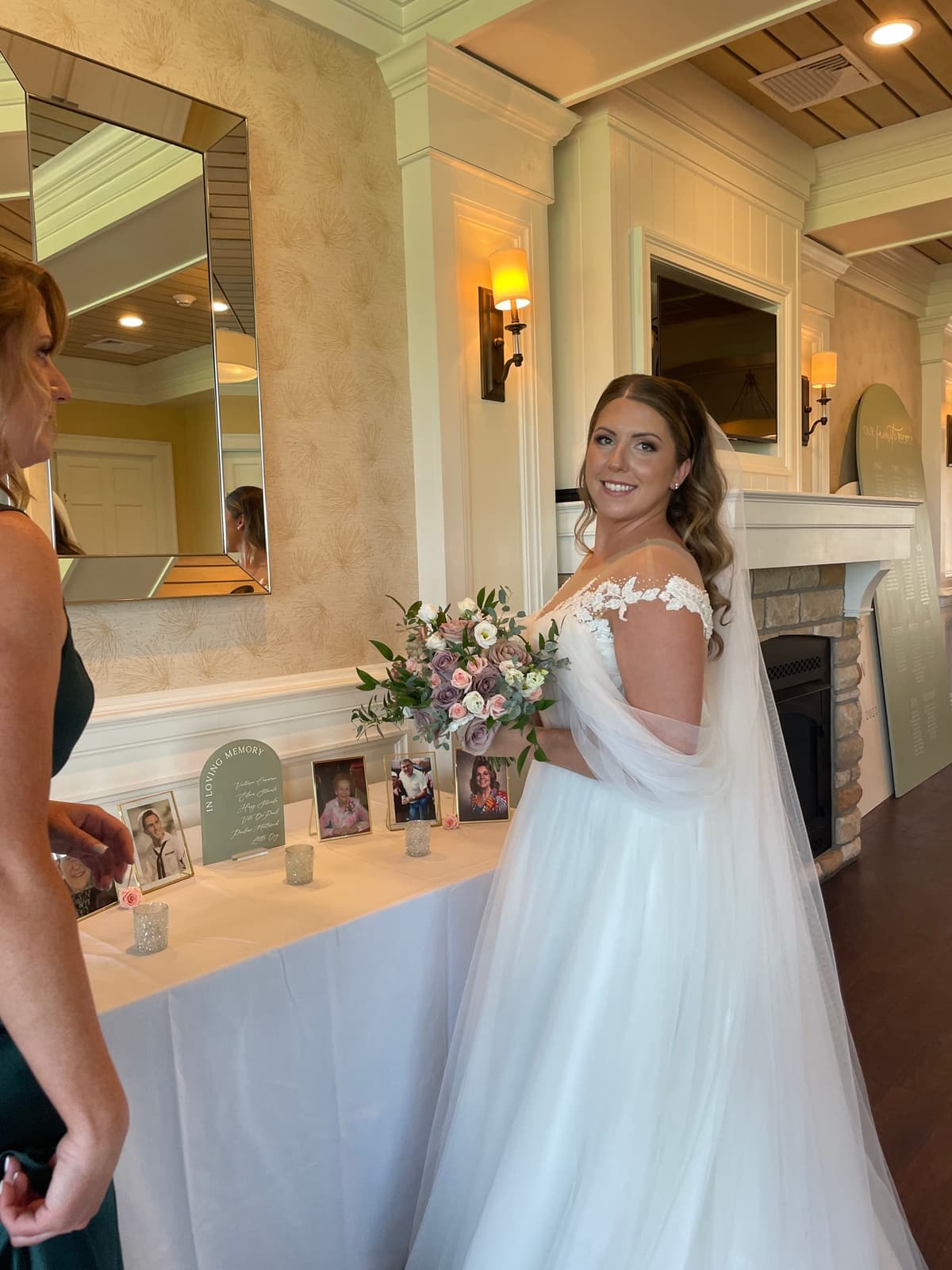 A smiling bride in a white lace wedding dress holds a round bouquet of dusty mauve and blush roses with eucalyptus greenery, standing next to a memorial table with framed photos.