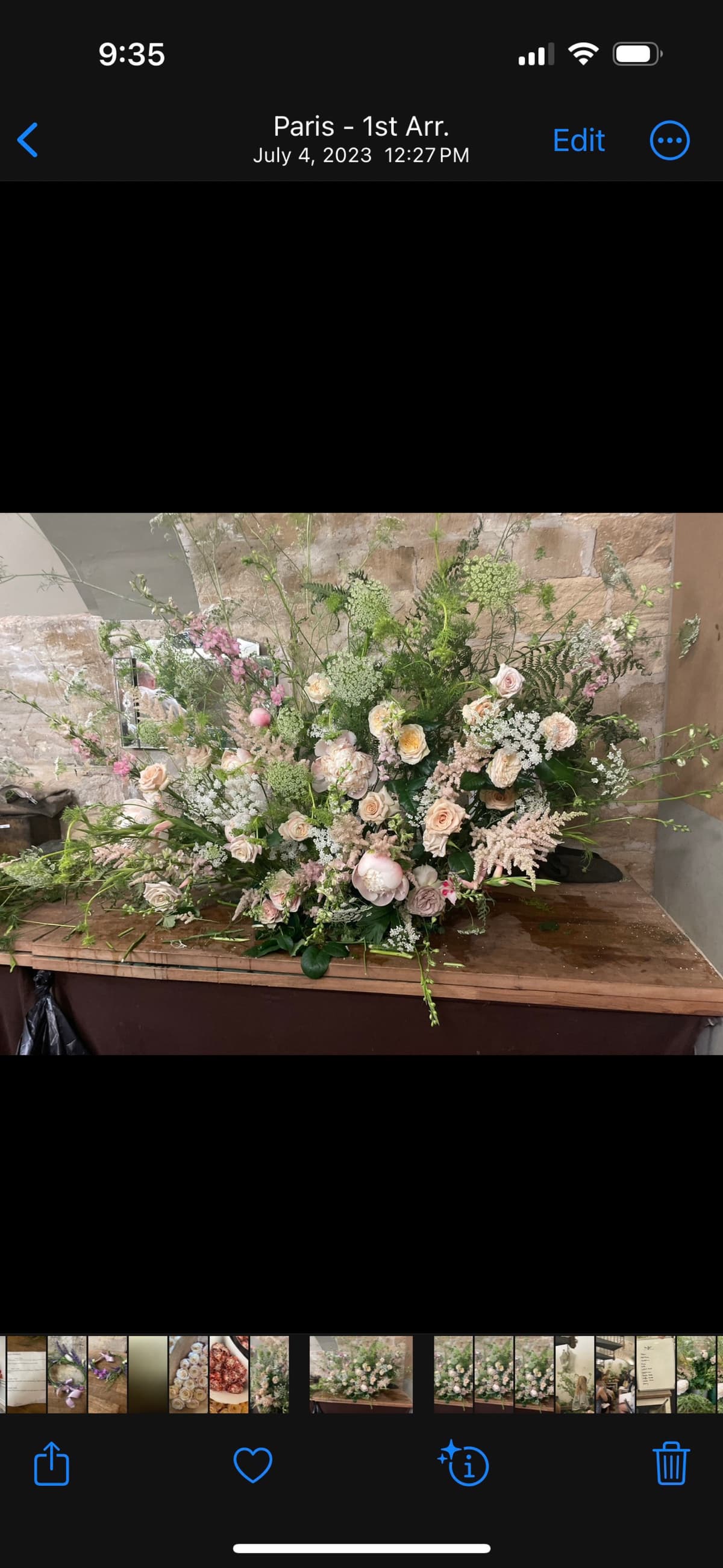 A large, lush floral arrangement on a wooden table featuring blush garden roses, pink peonies, and tall stems of Queen Anne's lace and greenery against a stone wall background.