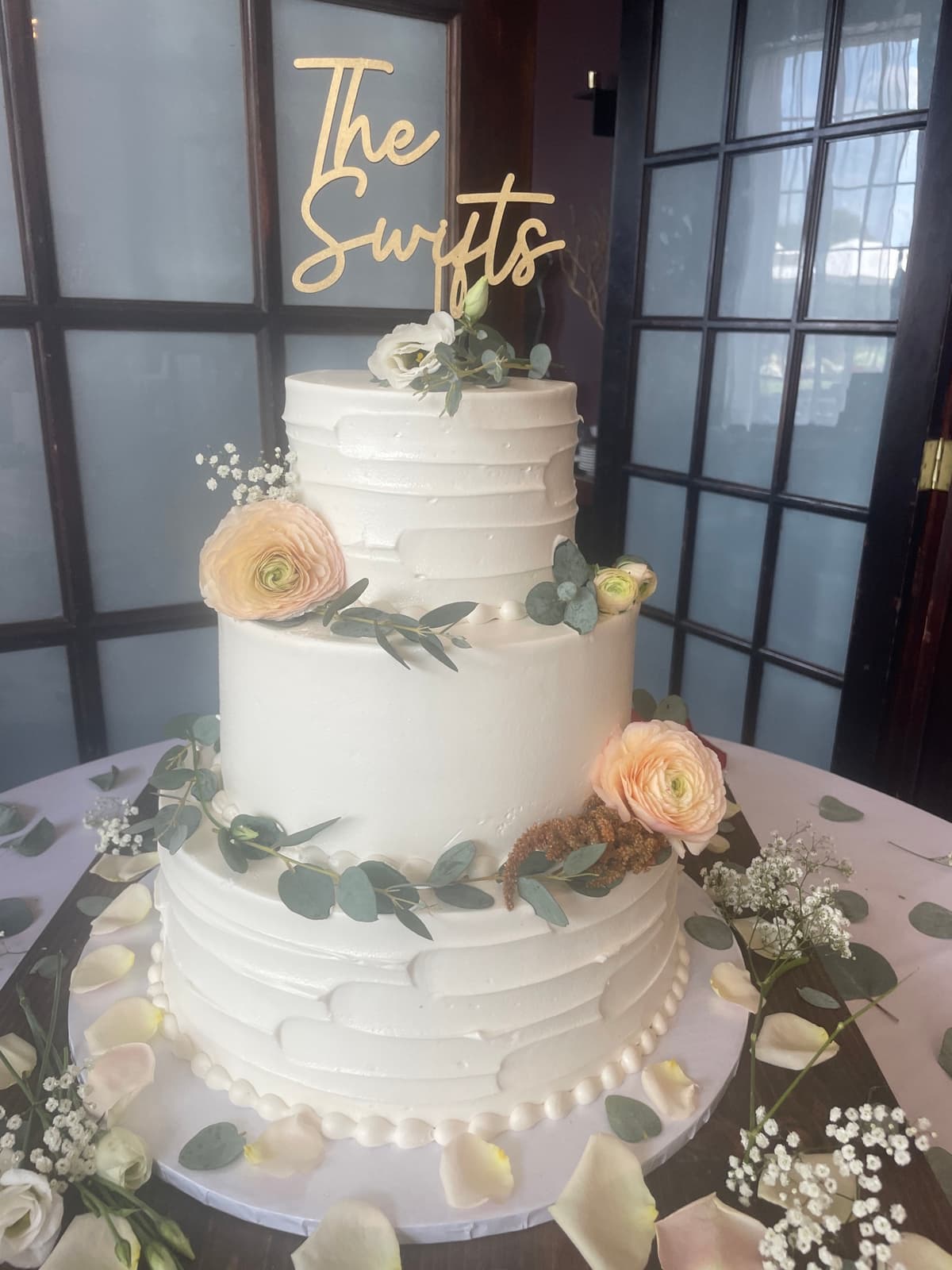 A two-tier white wedding cake featuring a gold topper reading 'The Swifts', decorated with cascading fresh peach ranunculus, eucalyptus greenery, and sprigs of baby's breath.