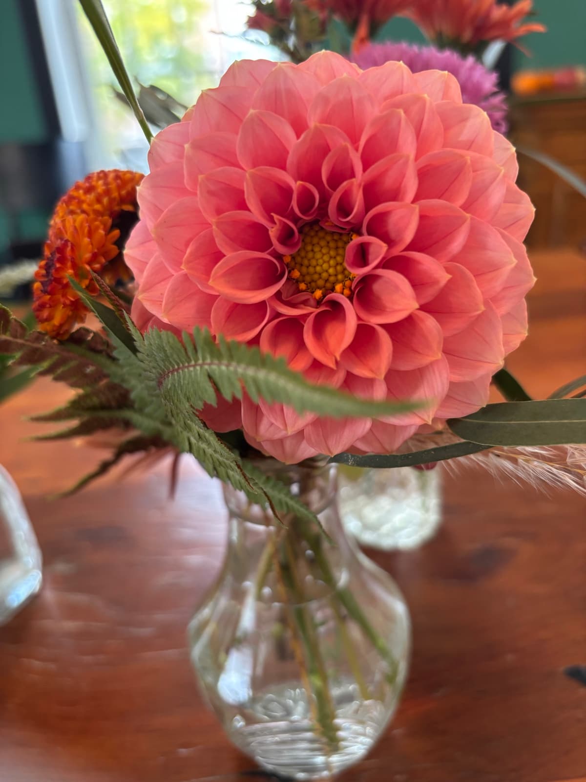 Top-down close-up of a large coral-pink dahlia in a clear cut-glass vase on a wooden table, accented by a green fern frond and a small orange flower.