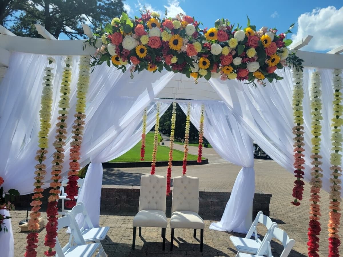 A white wedding pergola draped in sheer fabric, featuring a top arrangement of sunflowers and coral roses, with vertical floral garlands hanging in an ombré gradient from white to burgundy behind two tufted chairs.