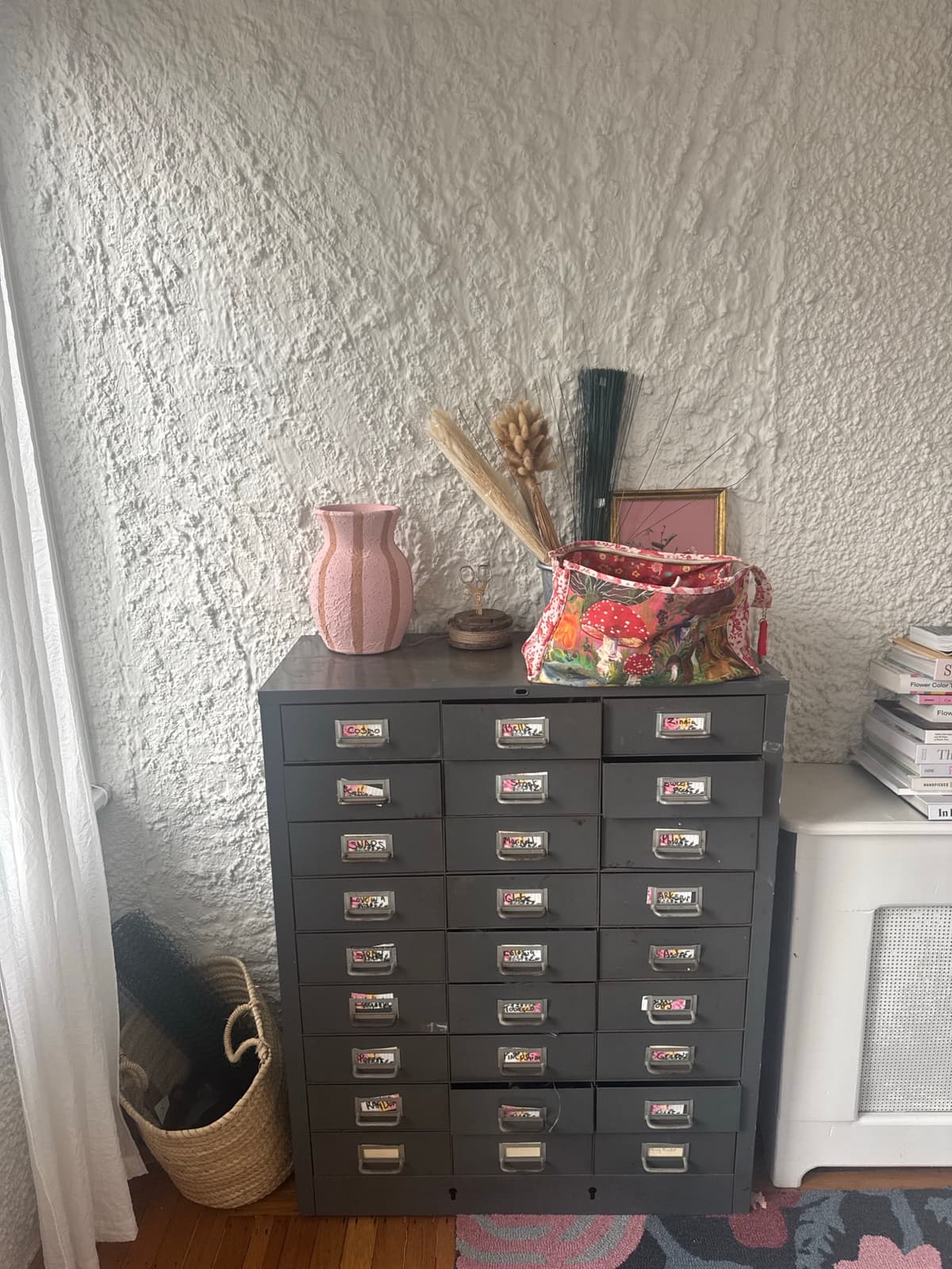 A dark grey industrial multi-drawer cabinet labeled with flower names stands against a textured white wall, styled with a pink striped vase, a floral bag, and a bundle of dried grasses.