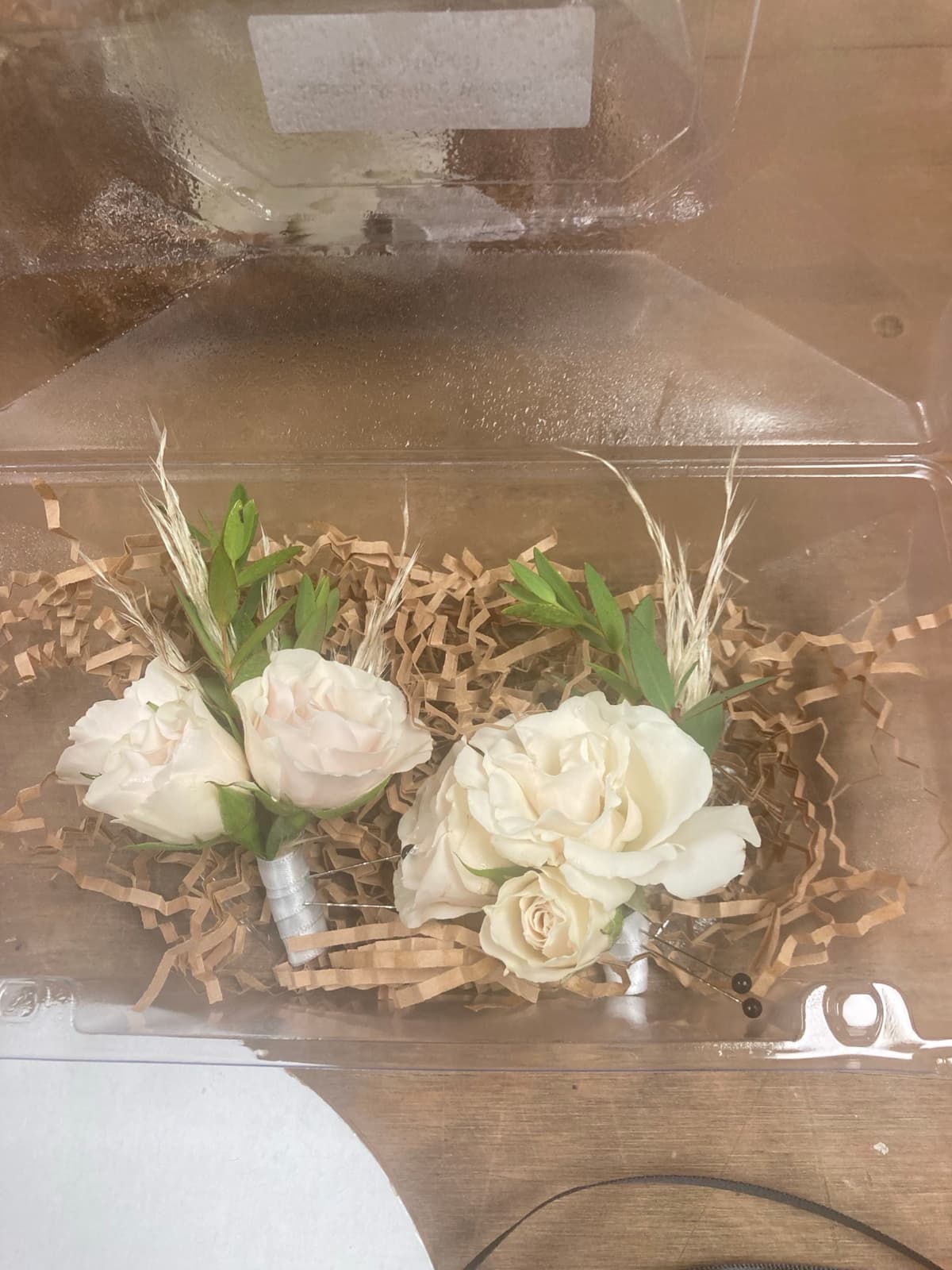 Two creamy white rose boutonnieres with dried grass accents resting on brown crinkle paper inside a clear plastic container.