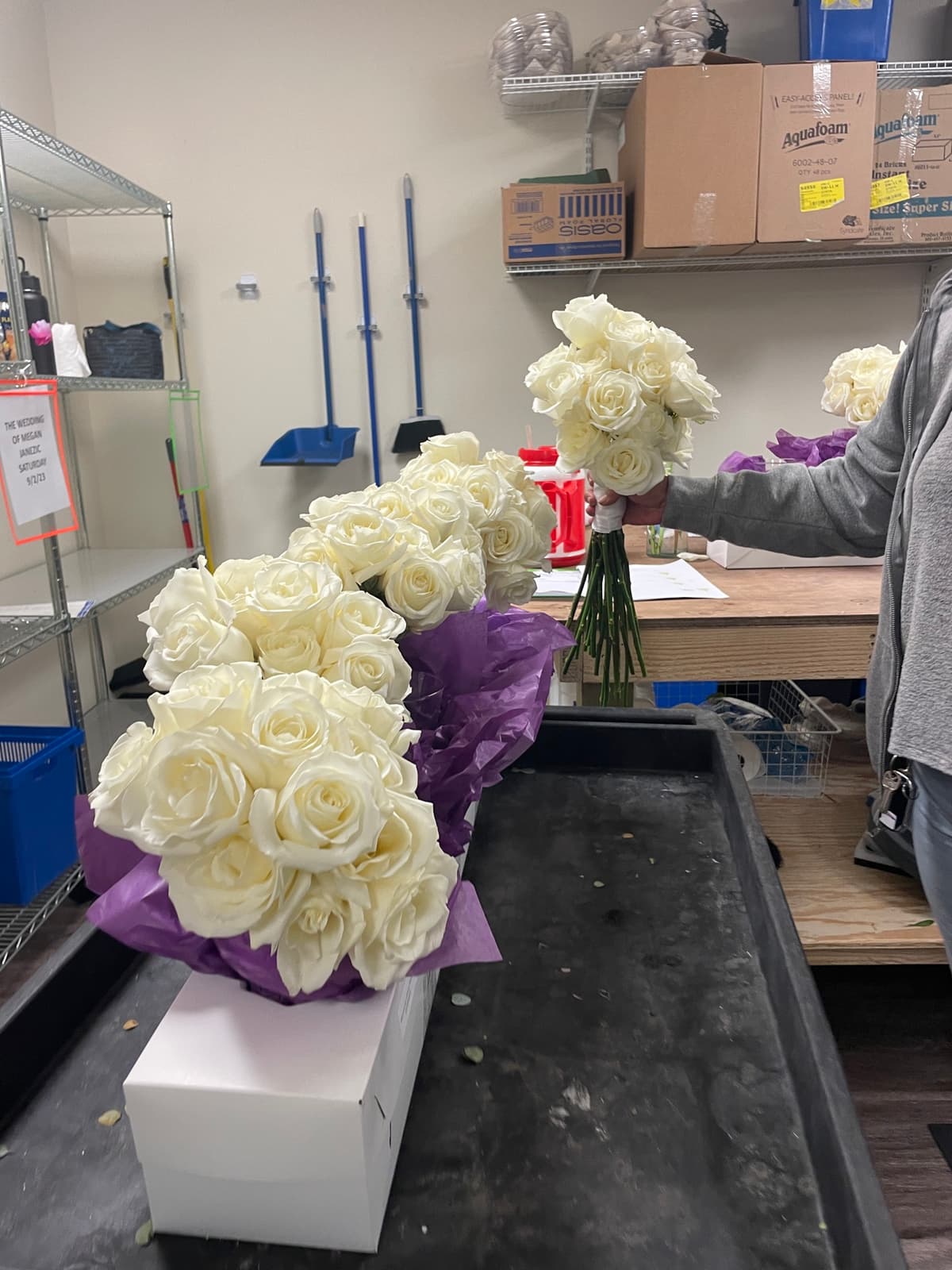 A florist holding a hand-tied bouquet of creamy white roses in a workshop setting, with a larger cluster of white roses resting on purple tissue paper nearby.