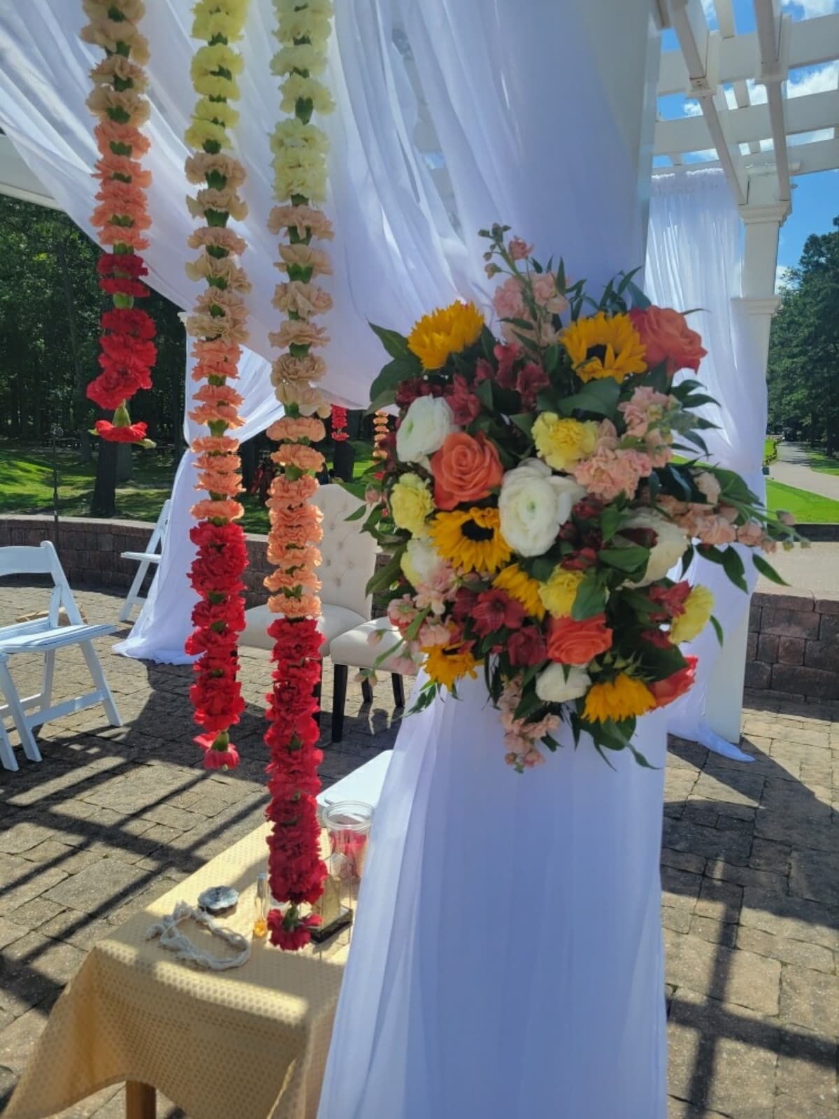 A sunny outdoor wedding ceremony setup featuring white fabric draping over a pergola. Three vertical floral garlands hang on the left, displaying an ombre gradient from yellow to red. On the right pillar, a large floral spray features sunflowers, white ranunculus, and coral roses.