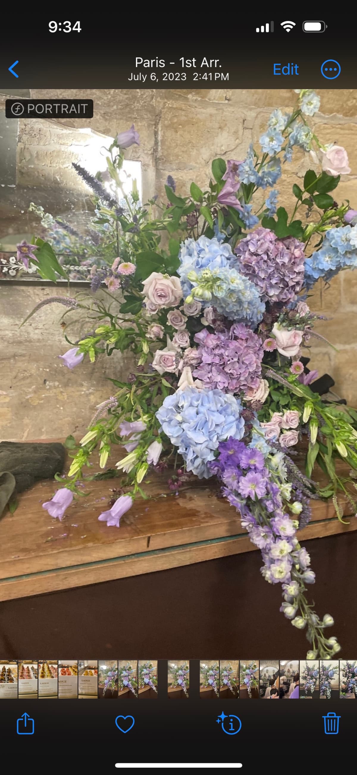 A lush, cascading floral arrangement featuring blue and purple hydrangeas, tall delphiniums, and pale lavender roses set against a rustic stone wall.