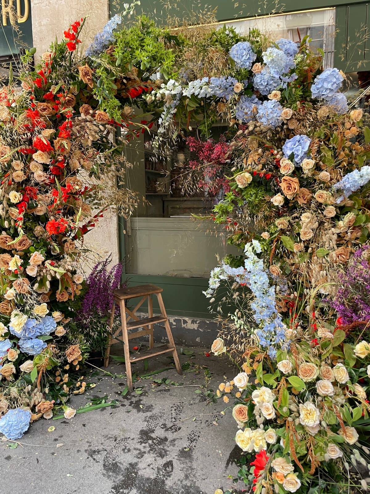 A lush, asymmetrical floral archway framing a storefront window and door. The installation features blue hydrangeas, sandy-colored roses, tall blue delphiniums, and dried grasses. A small wooden stool sits in the center of the frame.