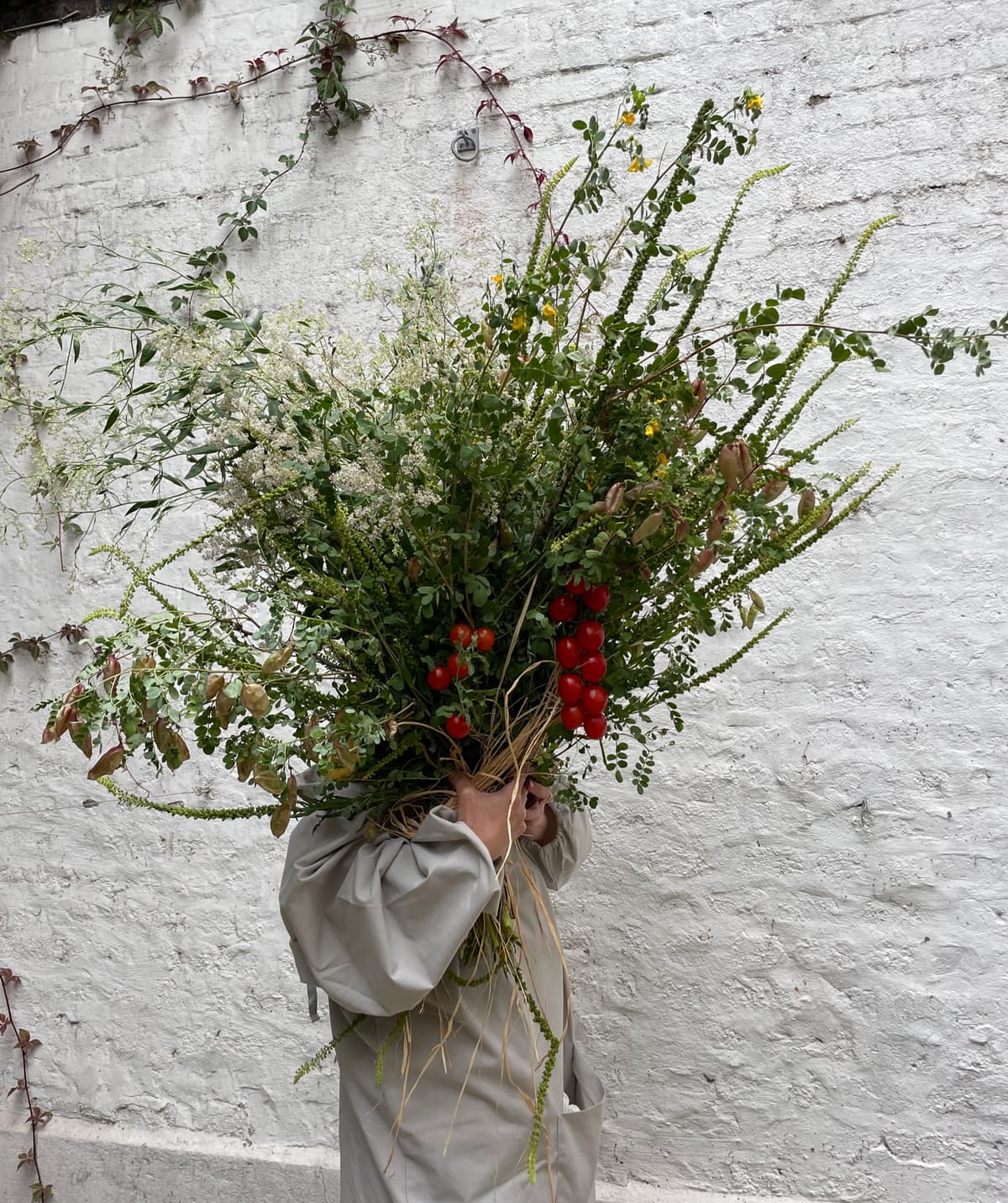 A person wearing a beige smock holds a massive, overflowing floral arrangement filled with wild greens, white wildflowers, and red cherry tomatoes on the vine against a white brick wall.